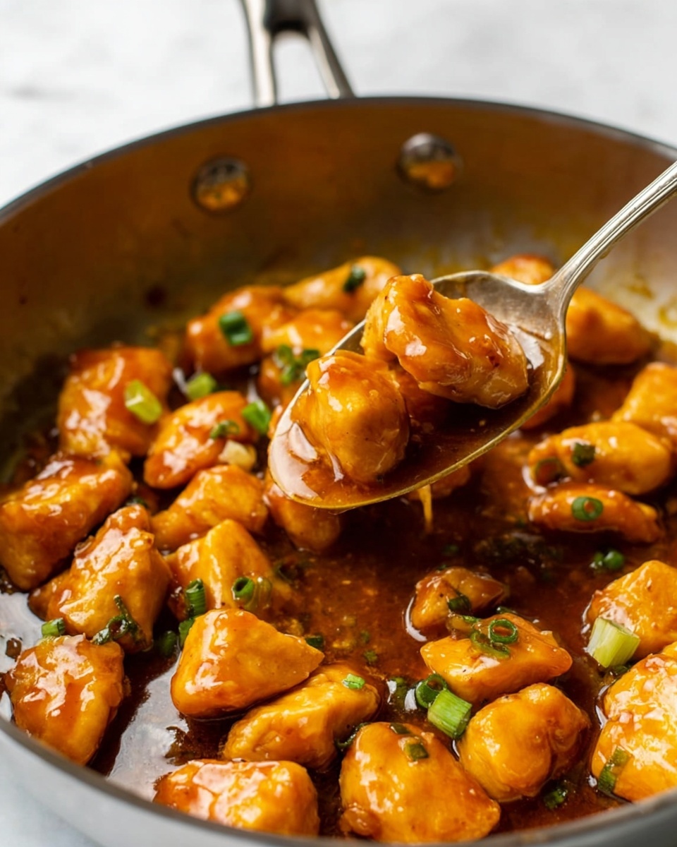 The image shows small golden brown pieces of cooked chicken coated in a shiny, thick orange-brown sauce inside a pan. The chicken pieces are irregular in shape and have a slightly crispy texture underneath the sauce. Bright green chopped scallions are scattered on top, adding a fresh color contrast. A spoon is lifting two pieces of chicken out of the sauce, with some sauce dripping back into the pan. The background is a white marbled surface. photo taken with an iphone --ar 4:5 --v 7