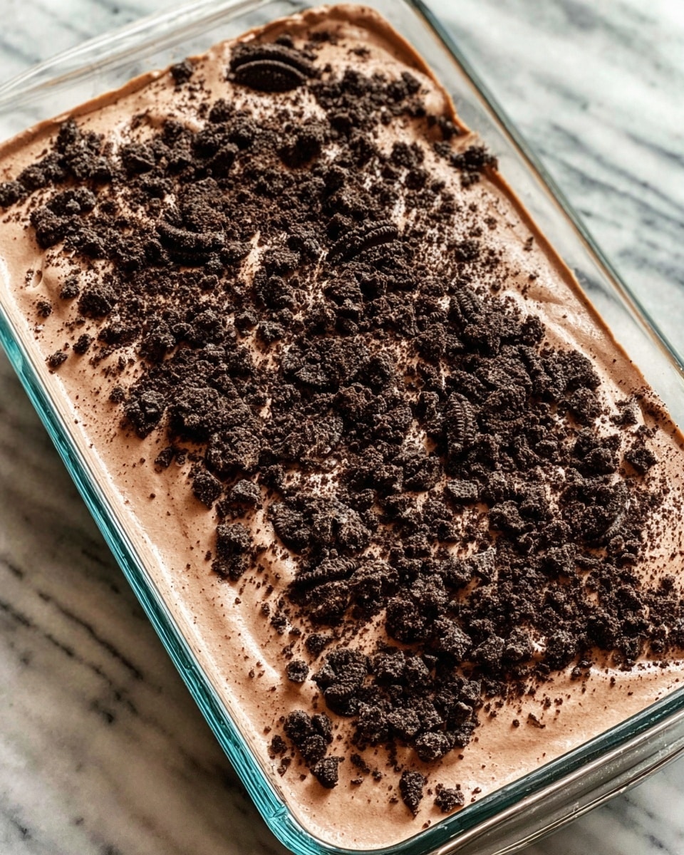 A rectangular glass dish filled with one thick, smooth layer of light brown chocolate mousse, topped with a generous amount of crushed dark chocolate cookies spread unevenly across the surface, creating a textured contrast. The dish is placed on a white marbled surface. photo taken with an iphone --ar 4:5 --v 7