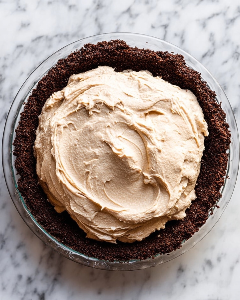 The image shows a clear glass pie dish filled with a dark brown, crumbly crust that forms the bottom and sides. On top of this crust is a large dollop of light brown, creamy filling with a smooth and swirled texture, placed mostly in the center. The pie dish sits on a white marbled surface. photo taken with an iphone --ar 4:5 --v 7