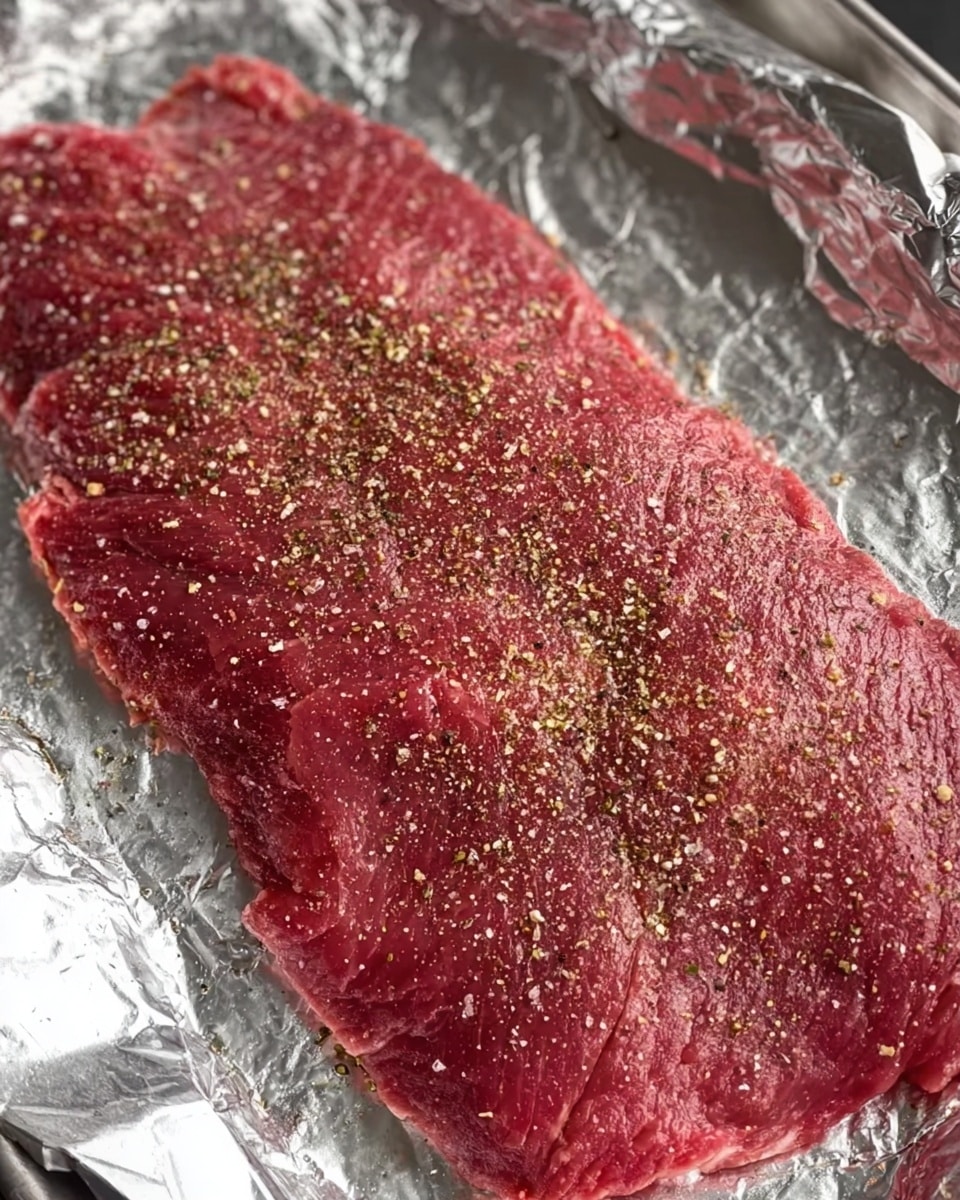 The image shows a large thin piece of raw red meat laid flat on a sheet of shiny aluminum foil that covers a metal tray. The meat is evenly sprinkled with coarse black pepper and light tan dried herbs, giving a speckled texture on the bright red surface. The edges of the meat are irregular, showing natural muscle lines and slight marbling. The aluminum foil wrinkles slightly around the meat, reflecting some light, and the background beneath the tray is mostly out of focus with a white marbled texture. photo taken with an iphone --ar 4:5 --v 7