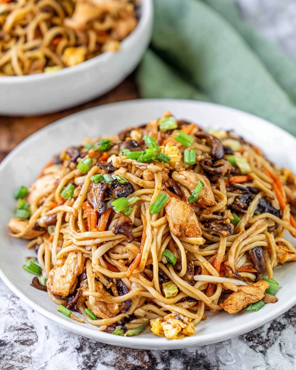 A white plate holds a serving of stir-fried noodles mixed with light and golden brown chicken pieces, thin orange carrot strips, dark brown mushrooms, and small bits of scrambled egg. The noodles are light tan and slightly shiny, layered with pieces of green onion scattered on top and throughout the dish. The plate is placed on a surface that looks like white marble with gray veins, with a soft green cloth blurred in the background. There is a white bowl filled with more of the same noodles partially visible behind the plate. photo taken with an iphone --ar 4:5 --v 7