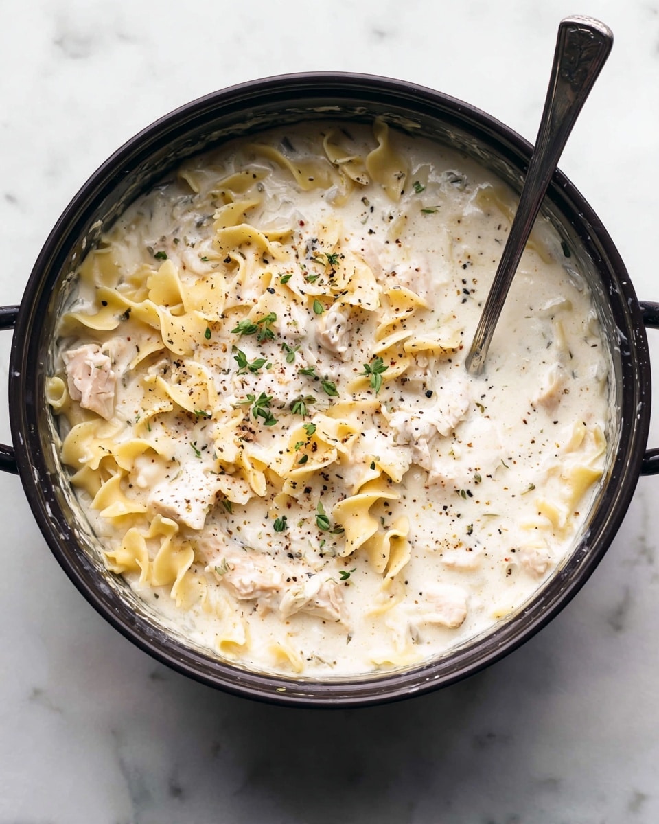 A black pot filled with creamy white sauce pasta mixed with yellow, curly-edged lasagna noodles and pieces of pale pink chicken. The sauce looks smooth and thick, speckled with black pepper and small sprigs of green herbs sitting on top. A metal spoon rests inside the pot. The pot is placed on a white marbled surface. Photo taken with an iphone --ar 4:5 --v 7