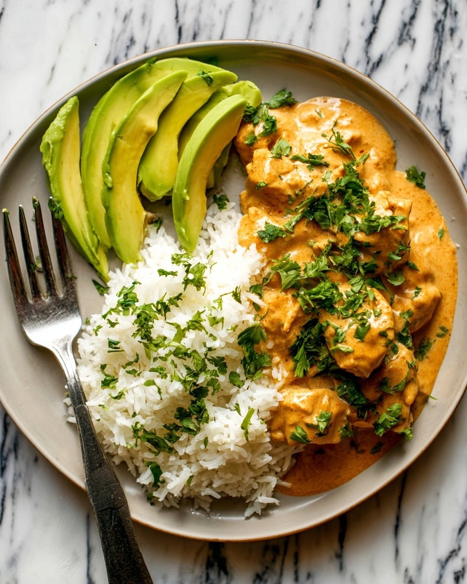 A white plate holds a base layer of soft white rice with visible grains. On top, several pieces of meat are covered in thick, creamy orange sauce with small bits of herbs and spices. Fresh green parsley leaves are sprinkled over the sauce, adding contrast. In a corner, thin slices of light green avocado rest against the edge of the plate. The dish is set on a white marbled surface. photo taken with an iphone --ar 4:5 --v 7