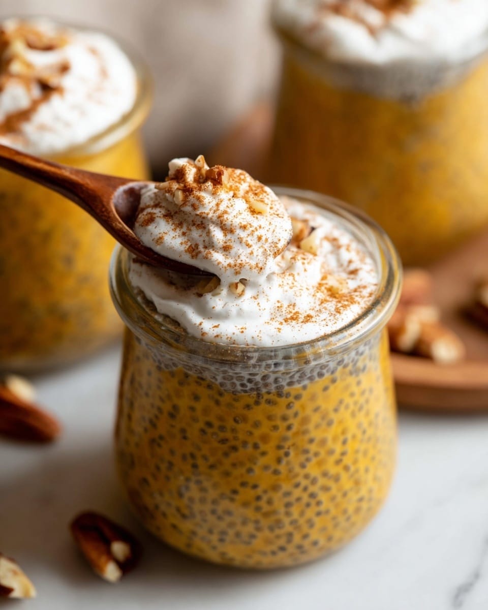 A close-up of a small clear glass jar filled with three visible layers: the bottom and main layer is a rich golden yellow pudding with visible black chia seeds speckled throughout, giving it a textured look; on top of this is a dollop of white whipped cream with a smooth and fluffy texture, sprinkled lightly with a brown powder, likely cinnamon; a wooden spoon inside the jar lifts a scoop showing the thick pudding and cream mixed with small brown nut pieces. The jar sits on a white marbled surface with parts of other similar jars and scattered nuts in the blurry background. Photo taken with an iphone --ar 4:5 --v 7