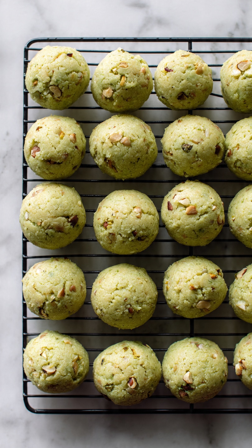 The image shows small, light green cookie-like dough balls with bits of nuts or seeds mixed inside. They are arranged in rows on a black wire cooling rack. The dough balls have a rough and bumpy texture, with some slightly browned spots on top. The baking rack is placed on a white marbled surface. The cookies look fresh and ready to bake or cool. photo taken with an iphone --ar 4:5 --v 7