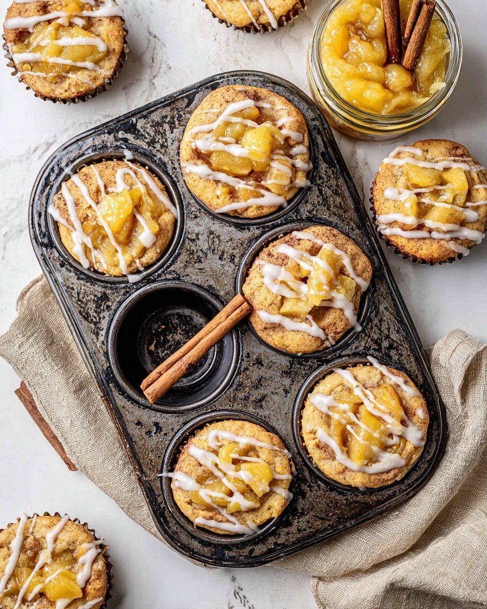The image shows a dark, worn muffin tin holding six small round pastries, each with two layers—a light golden-brown base and a topping of chunky yellow apple filling with a shiny texture. The top layer is finished with white icing drizzled in thin lines across the pastries. Two cinnamon sticks rest in one empty muffin cup. Around the muffin tin on a white marbled surface are more pastries topped with the same apple filling and white icing, alongside a jar filled with the same yellow apple filling and topped with a cinnamon stick. A beige linen cloth is partly under the muffin tin. Photo taken with an iphone --ar 4:5 --v 7