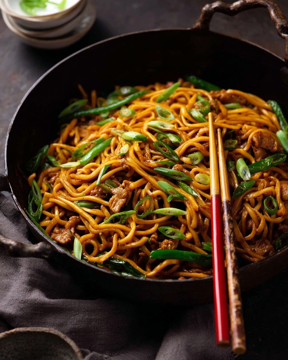 A dark pan filled with orange-brown noodles mixed with small pieces of light brown cooked meat and bright green vegetable strips, topped with thin sliced green onions scattered across the dish; a pair of red and light beige chopsticks rest inside the pan on the right side, gripping some noodles. The pan is placed on a dark surface with a folded gray cloth nearby, and stacked white bowls are blurred in the background. photo taken with an iphone --ar 4:5 --v 7