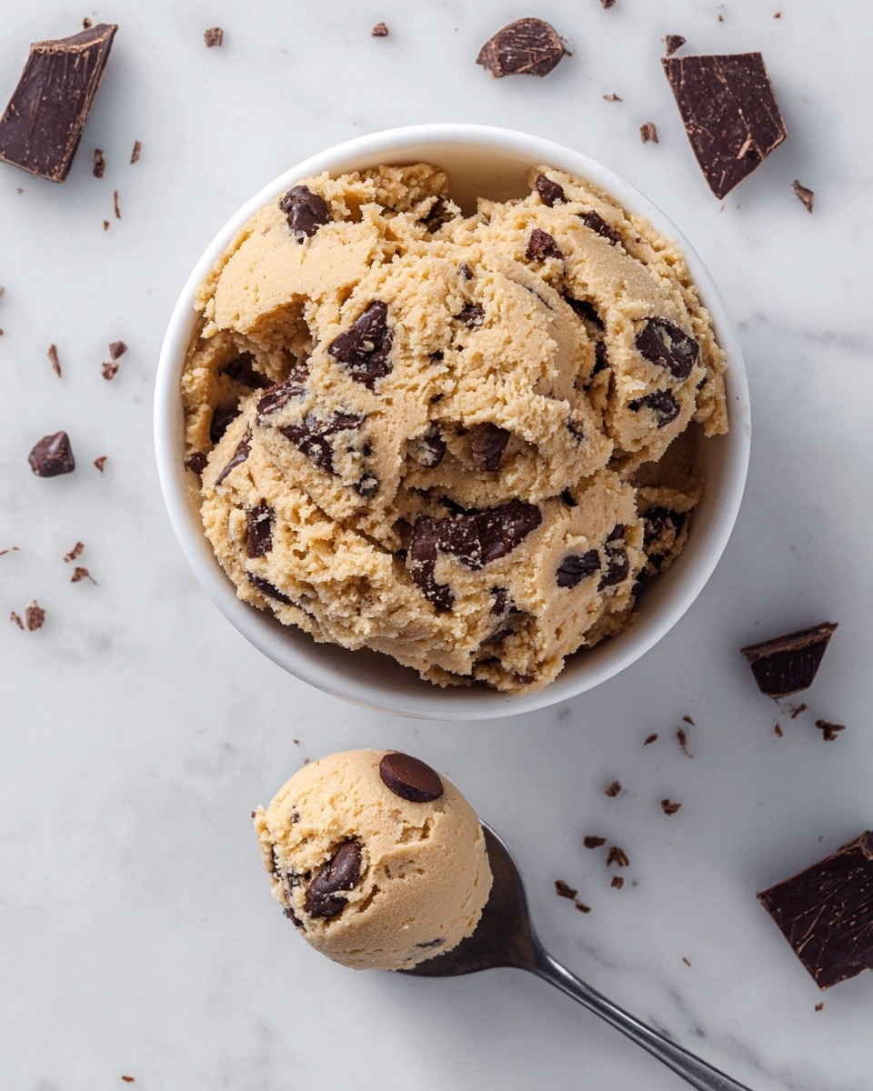 A white bowl filled with thick, light brown cookie dough dotted with dark brown chocolate chunks is placed on a white marbled surface. Scattered around the bowl are small pieces of broken chocolate. In front of the bowl, a spoon holds a rounded scoop of the same cookie dough, showing its soft, grainy texture and embedded chocolate chunks. The spoon is metallic and lies directly on the marbled surface. The image has a clean, simple style with a top-down view. photo taken with an iphone --ar 4:5 --v 7