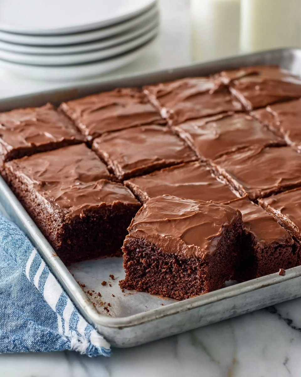 The image shows a rectangular metal tray holding a chocolate cake with one square piece removed from the bottom left corner. The cake has two visible layers: a moist, dark brown chocolate cake base and a smooth, shiny, thick chocolate frosting top layer with slight cracks on its surface. The cake is cut into a grid of rectangular sections. The tray is placed on a white marbled surface with a blue and white striped cloth partially visible on the lower left. In the background, there is a blurry stack of white bowls and a glass of milk. photo taken with an iphone --ar 4:5 --v 7