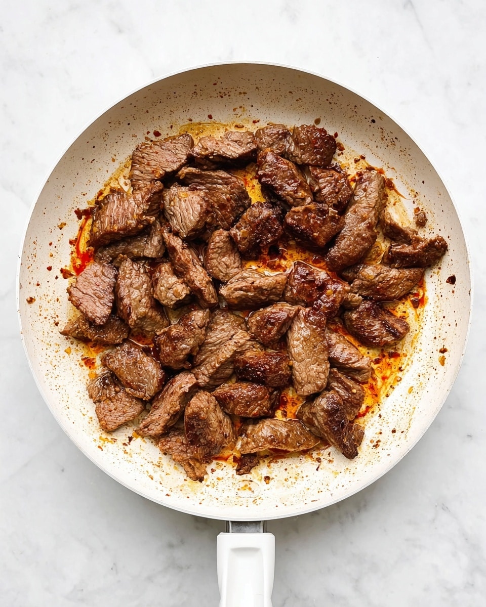 The image shows a white frying pan on a white marbled surface filled with many pieces of cooked beef. The beef pieces are brown with slightly darker edges and some caramelized spots showing rich browning. The pan has reddish-brown oil and seasoning marks scattered around the beef, adding a textured look. The beef pieces vary in size and are spread out evenly, creating a layer of cooked meat that fills most of the pan's surface. photo taken with an iphone --ar 4:5 --v 7