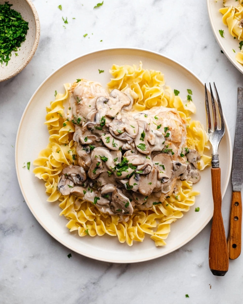 A white round plate holds three layers: the bottom layer is a bed of yellow curly egg noodles spread evenly, the middle layer is a large piece of light brown cooked chicken, and the top layer is a thick cream sauce with light brown cooked mushroom slices scattered on the chicken and noodles, garnished with small green parsley bits. The plate is on a white marbled surface, with a knife and fork to the right with wooden handles, and a small bowl with green herbs partially visible to the left. Photo taken with an iphone --ar 4:5 --v 7