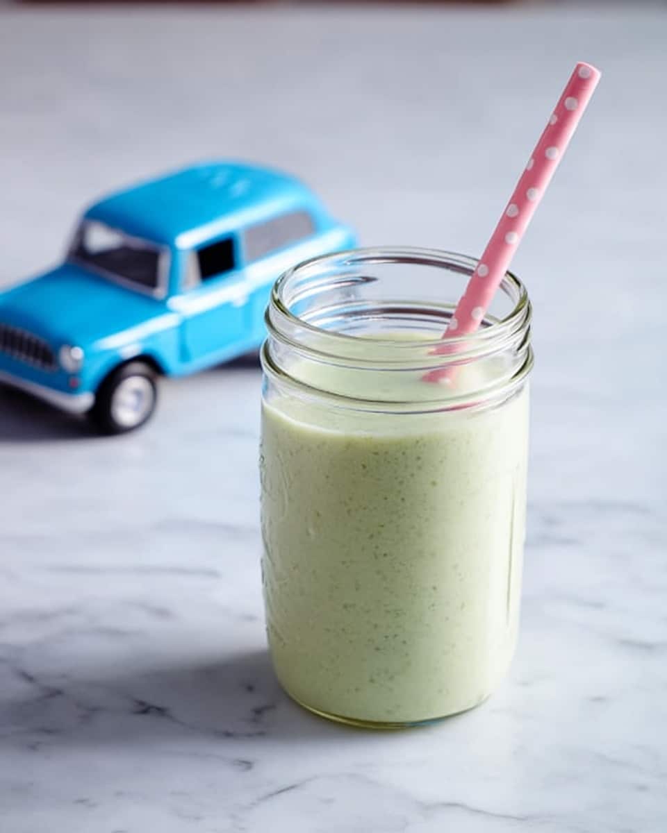 The image shows a clear glass jar filled with a smooth, light green smoothie. The smoothie has a creamy texture and fills the jar almost to the top. A blue and white striped straw is placed inside the jar, leaning slightly to the right. The jar sits on a white marbled surface with a soft gray cloth folded behind it. Two light green fruit cubes are placed near the bottom right corner of the jar. The background has a rustic wooden look in faded gray tones. Photo taken with an iphone --ar 4:5 --v 7