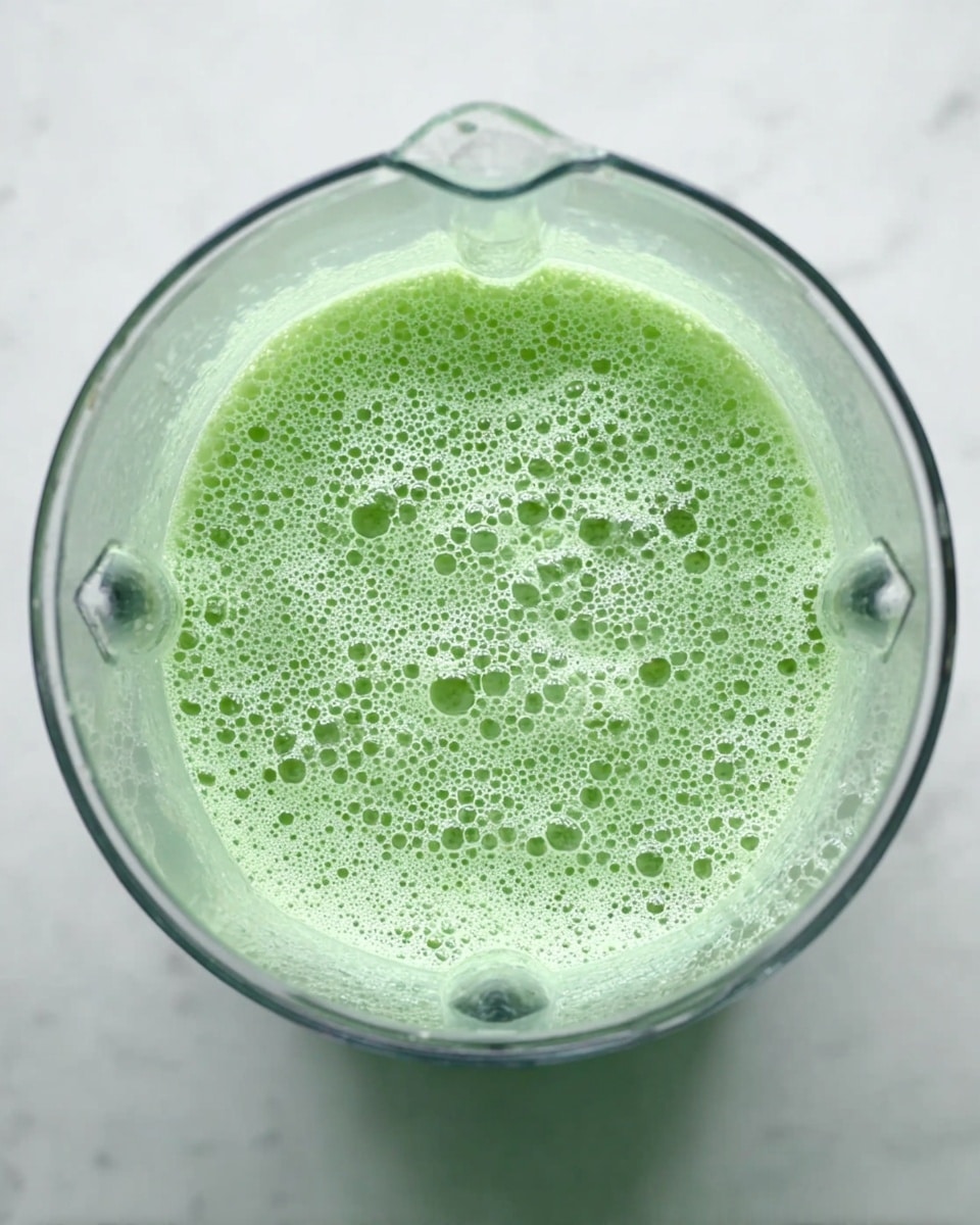 The image shows a clear blender container filled with a light green liquid that is frothy and bubbly on the surface. The bubbles are small to medium size, covering most of the liquid's top layer. The blender is set on a white marbled surface, and the photo is taken from directly above, focusing on the texture of the green mixture inside the blender. photo taken with an iphone --ar 4:5 --v 7