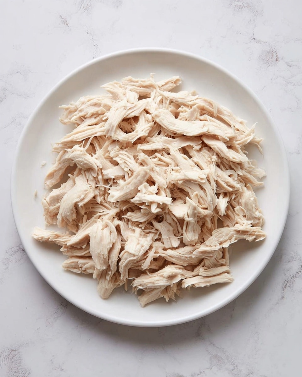 A white plate filled with a single layer of shredded light beige cooked chicken pieces, spread evenly and loosely across the plate. The chicken strands are fibrous and soft, with varying lengths and thicknesses, showing natural texture and slight curve. The plate sits on a white marbled surface with subtle gray veins, creating a clean and simple background. Photo taken with an iphone --ar 4:5 --v 7