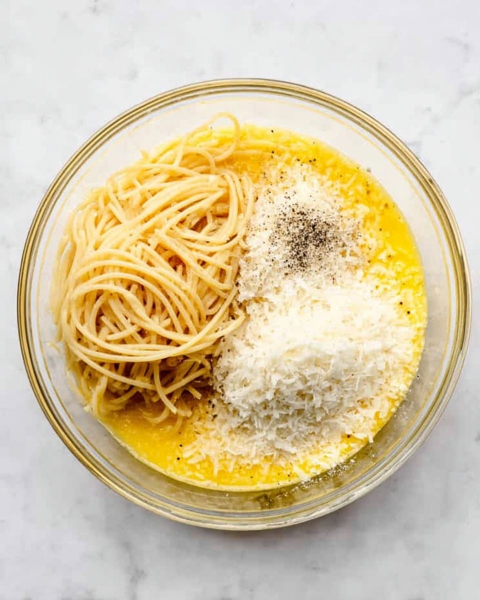 A clear glass bowl sits on a white marbled surface, filled with three main layers visible from above. At the bottom, there is a layer of yellow liquid, likely egg yolk or sauce. On top of this, a bundle of light beige spaghetti noodles lies to one side. Around the noodles, there are three separate piles of finely grated white cheese, and a small amount of black pepper is sprinkled near the cheese. The colors contrast nicely with the clear bowl and the white marbled background, giving a simple but textured look. photo taken with an iphone --ar 4:5 --v 7