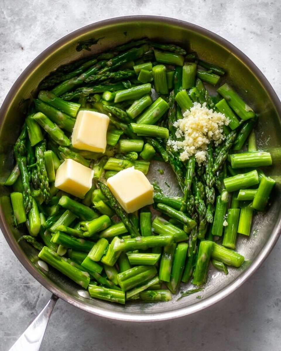 The image shows a round stainless steel pan filled with bright green asparagus pieces chopped into small sections. Two small light yellow butter chunks sit on top of the asparagus, along with a small pile of finely minced garlic near the center. The asparagus tips and stalks create a varied green texture throughout the pan. The pan rests on a white marbled surface, and the lighting highlights the fresh and ready-to-cook ingredients clearly. photo taken with an iphone --ar 4:5 --v 7