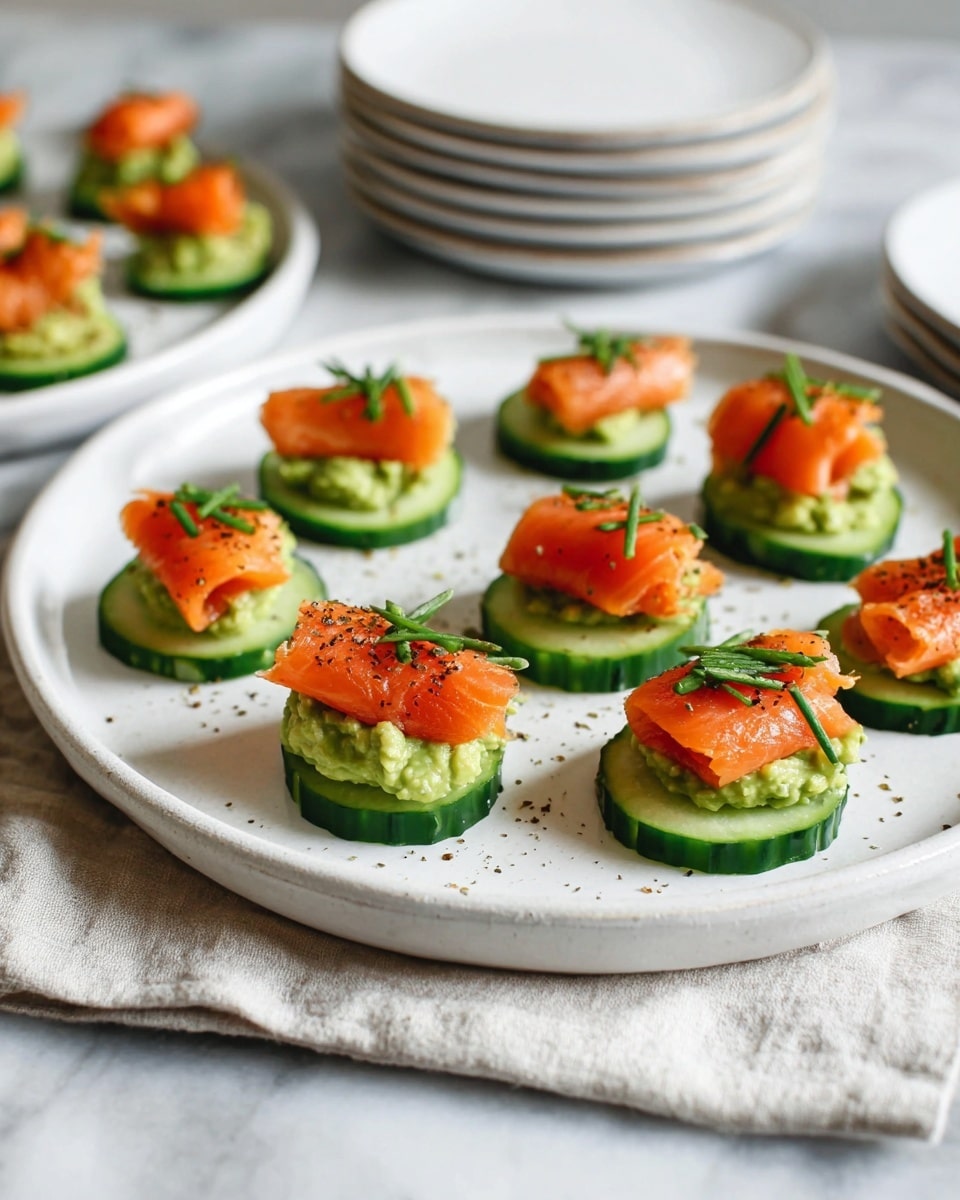 A white round plate holds eight cucumber slices arranged in a loose circle. Each cucumber slice has three layers: the bottom green cucumber slice with a dark green edge, a middle layer of light green mashed avocado spread roughly, and on top, bright orange smoked salmon pieces folded loosely. Small green chive pieces are scattered over each salmon portion, and a few black pepper specks are sprinkled across the plate. The plate rests on a white cloth over a white marbled surface. In the upper part, a smaller white round plate with two similar bites is partially visible. Photo taken with an iphone --ar 4:5 --v 7