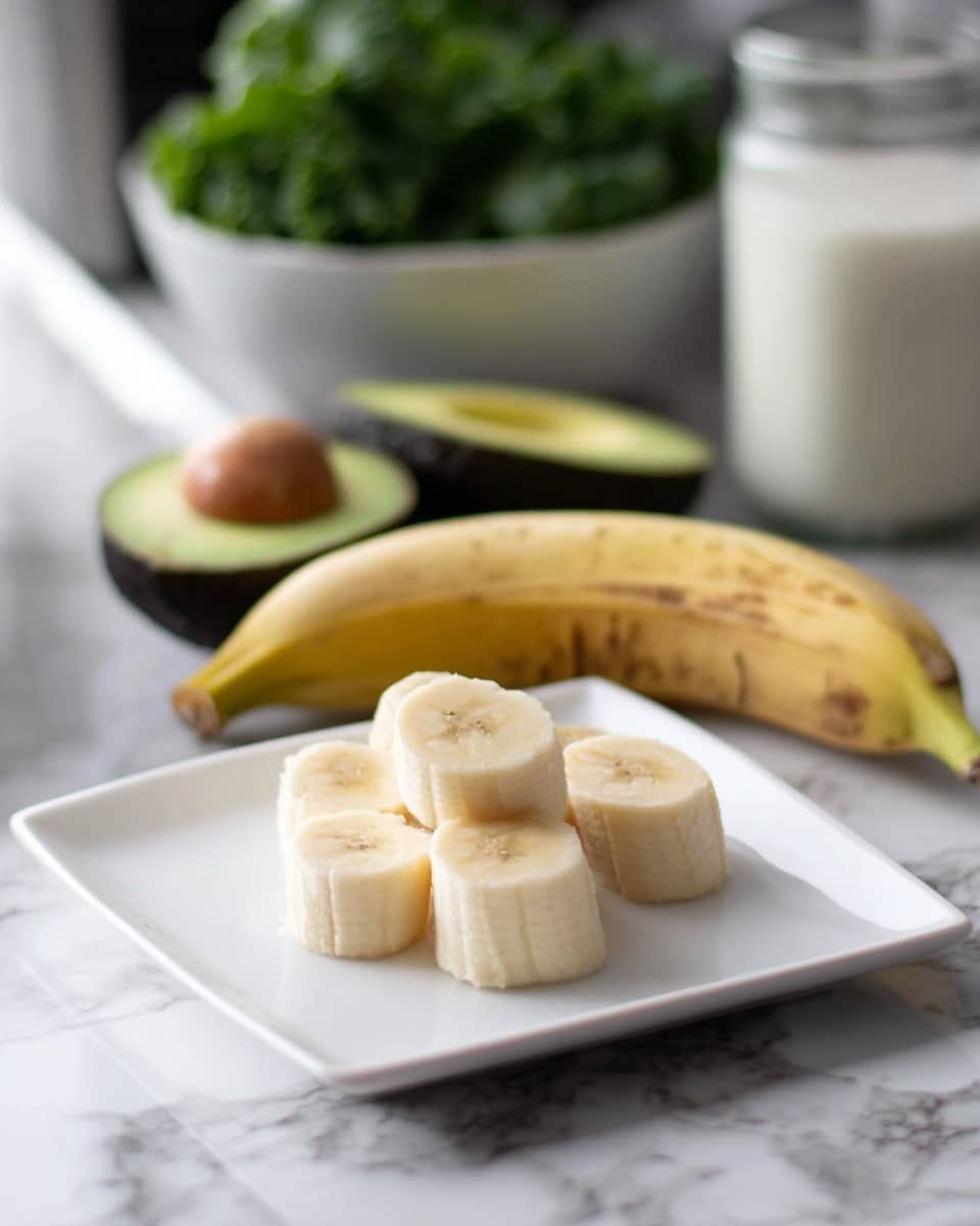 The image shows a white square plate with six pieces of peeled banana cut into short segments arranged in a small pile, showcasing their light creamy color and soft texture. Behind the peeled banana segments, there is one banana with yellow skin and brown spots laid horizontally on the plate. In the background, a halved avocado with green flesh and a large brown seed inside is visible next to a white bowl filled with leafy green vegetables, all set on a white marbled surface. There is also a glass jar filled with white liquid blurred in the back. The photo taken with an iphone --ar 4:5 --v 7