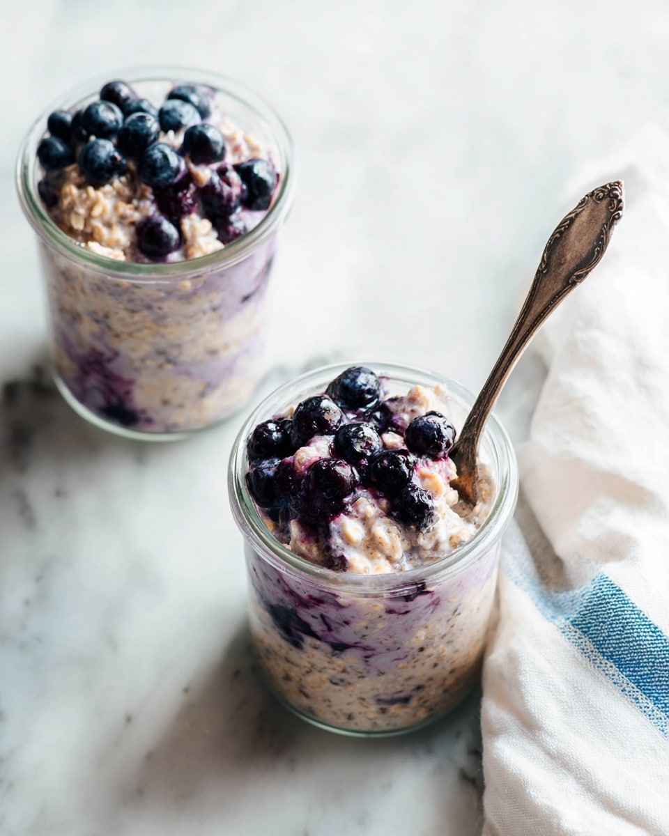 Two clear glass jars filled with a creamy oatmeal mixture with purple and white colors, topped with dark purple blueberries. The oatmeal has a thick texture with visible seeds and grains mixed in. One jar has a vintage silver spoon dipped inside, showing the chunky oatmeal and berries. The jars are placed on a white marbled surface with a white cloth with blue edges on the right side. The light is soft and natural, highlighting the smooth cream and shiny blueberries. photo taken with an iphone --ar 4:5 --v 7