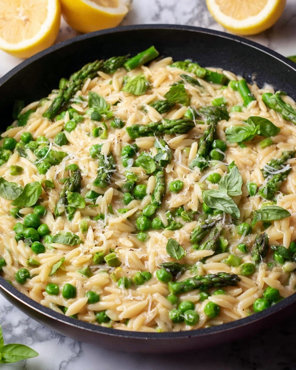 A close-up view of a round black pan filled with creamy orzo pasta mixed with bright green peas and chopped asparagus pieces, sprinkled with fresh green basil leaves and finely grated cheese on top. In the background, there are two halves of a lemon, showing their juicy yellow insides. The whole scene is set against a white marbled surface. Photo taken with an iphone --ar 4:5 --v 7