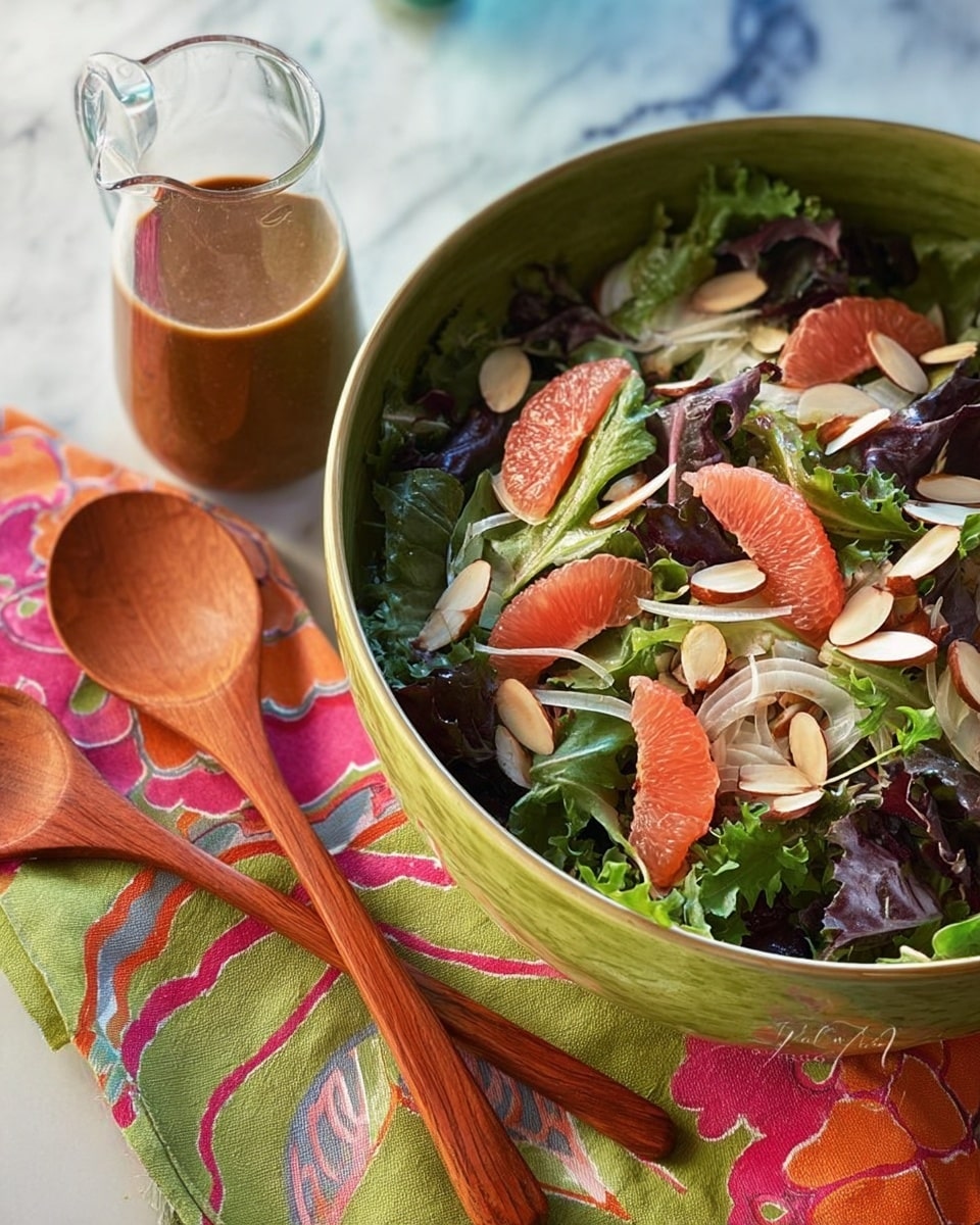 A large green bowl filled with a fresh mixed salad featuring dark green and purple leafy greens as the base layer, topped with peeled pink grapefruit segments, light green fennel slices, and whole almonds scattered throughout. Beside the bowl is a small glass pitcher filled with brown dressing, and next to it are two wooden salad spoons resting on a colorful patterned cloth with green, orange, and pink designs. The setting is on a white marbled surface with soft, natural lighting. Photo taken with an iphone --ar 4:5 --v 7