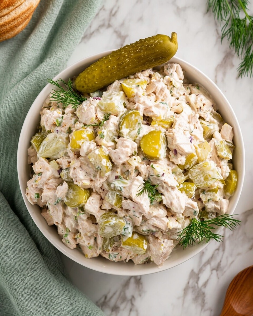 A white bowl filled with a creamy chicken salad made of small chunks of pale pinkish chicken mixed with light green celery pieces and finely chopped yellow pickles, all held together by a white creamy dressing. On top of the salad, there is a whole small green pickle and some fresh green dill leaves scattered around for garnish. The bowl sits on a white marbled surface with a soft green cloth partially visible on the left and a wooden spoon at the bottom right. photo taken with an iphone --ar 4:5 --v 7