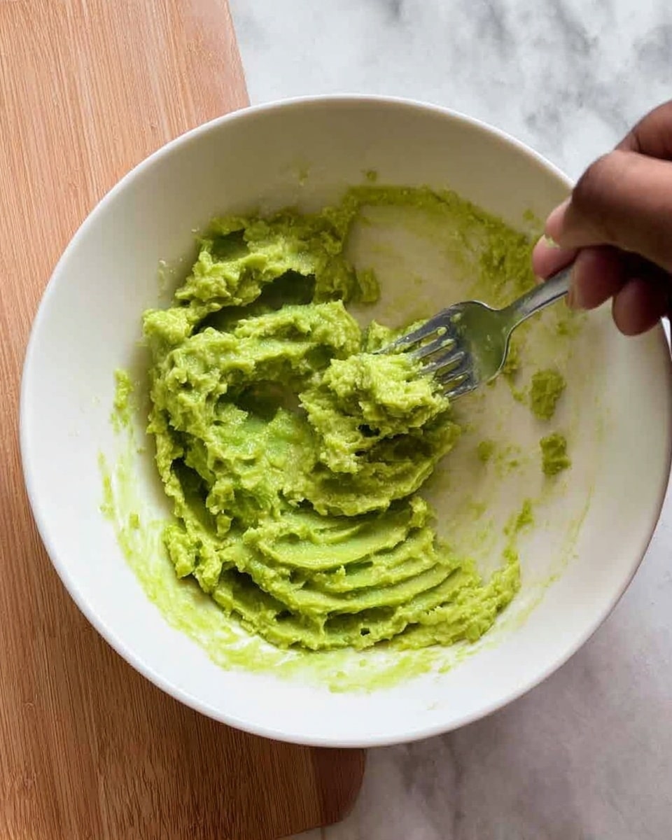 A white bowl filled with mashed green avocado, showing a slightly chunky texture with ridges and small pieces from being mashed with a silver fork. A woman's hand holding the fork is visible at the top right corner, pressing down into the avocado. The bowl sits on a wooden surface, and the background shows a white marbled texture. The avocado is spread across the bottom of the bowl with some bits smeared on the side. photo taken with an iphone --ar 4:5 --v 7