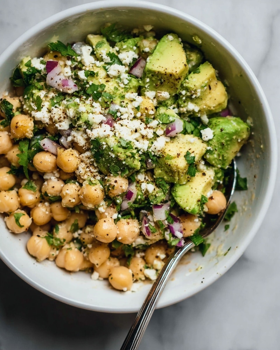 The image shows a white bowl filled with a salad made of three main layers: the bottom layer has beige chickpeas that are round and smooth, the middle layer shows green chunks of avocado with a soft and creamy texture, and the top layer is sprinkled with small bits of white cheese and finely chopped dark green cilantro. There are also small pieces of purple onion scattered throughout. A silver spoon rests inside the bowl on the right side, partially buried in the salad. The salad ingredients look fresh and lightly seasoned with black pepper visible on top. The bowl is placed on a white marbled surface. Photo taken with an iphone --ar 4:5 --v 7