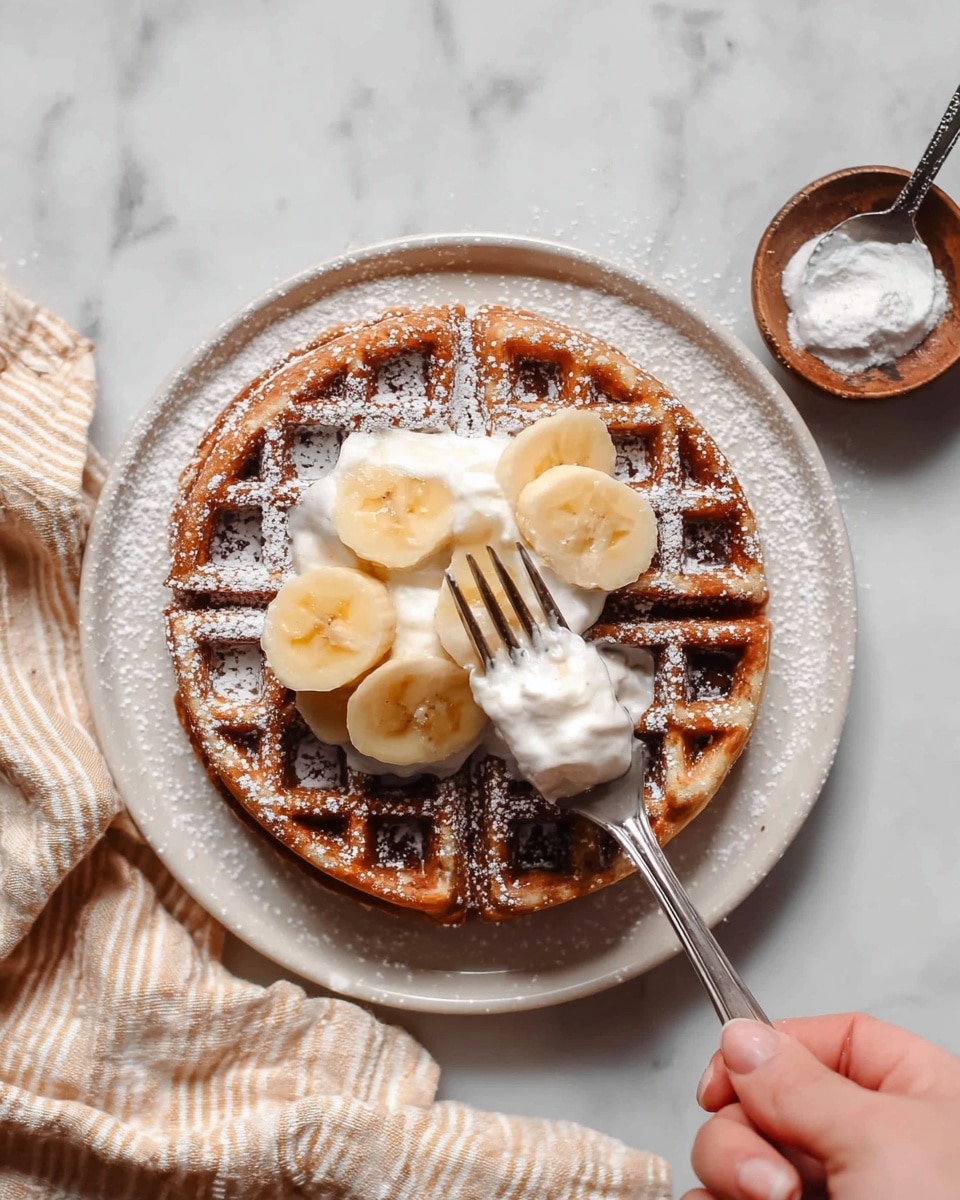 A round waffle with a golden brown color is placed on a white plate dusted with powdered sugar. The waffle is topped with a dollop of white cream and several slices of fresh yellow banana arranged on top. A fork is pressed into the waffle near the center while a spoon scoops some cream and banana slices. A woman's hand holds the fork. The plate sits on a white marbled surface, with a beige and white striped cloth partially visible beneath it. Nearby, a small wooden bowl holds more white powder with a metal spoon inside. Photo taken with an iphone --ar 4:5 --v 7