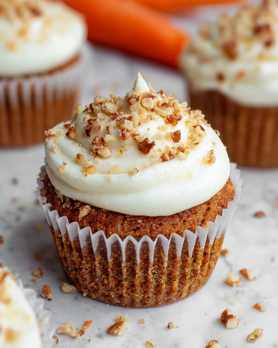A close-up of a carrot cupcake with a textured brown cake base held in a white paper liner, topped with a thick layer of smooth white frosting swirled on top, sprinkled with small chopped nuts in light brown and golden hues. The cupcake sits on a white marbled surface with some nut crumbs scattered around, and there are blurred similar cupcakes and an orange carrot in the soft background. Photo taken with an iphone --ar 4:5 --v 7