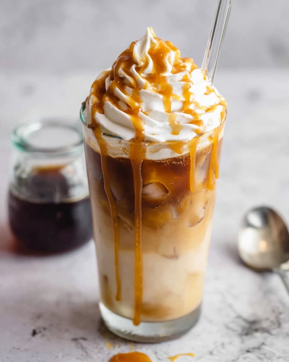 A tall clear glass filled with a light brown iced coffee layered with visible ice cubes inside, a dark syrup pooling at the bottom, topped with a thick layer of white whipped cream swirled in peaks, drizzled generously with golden caramel sauce over the whipped cream and sides of the glass, all placed on a white marbled surface with a small glass container of dark syrup and a silver spoon in the background, a clear straw inserted into the drink. photo taken with an iphone --ar 4:5 --v 7