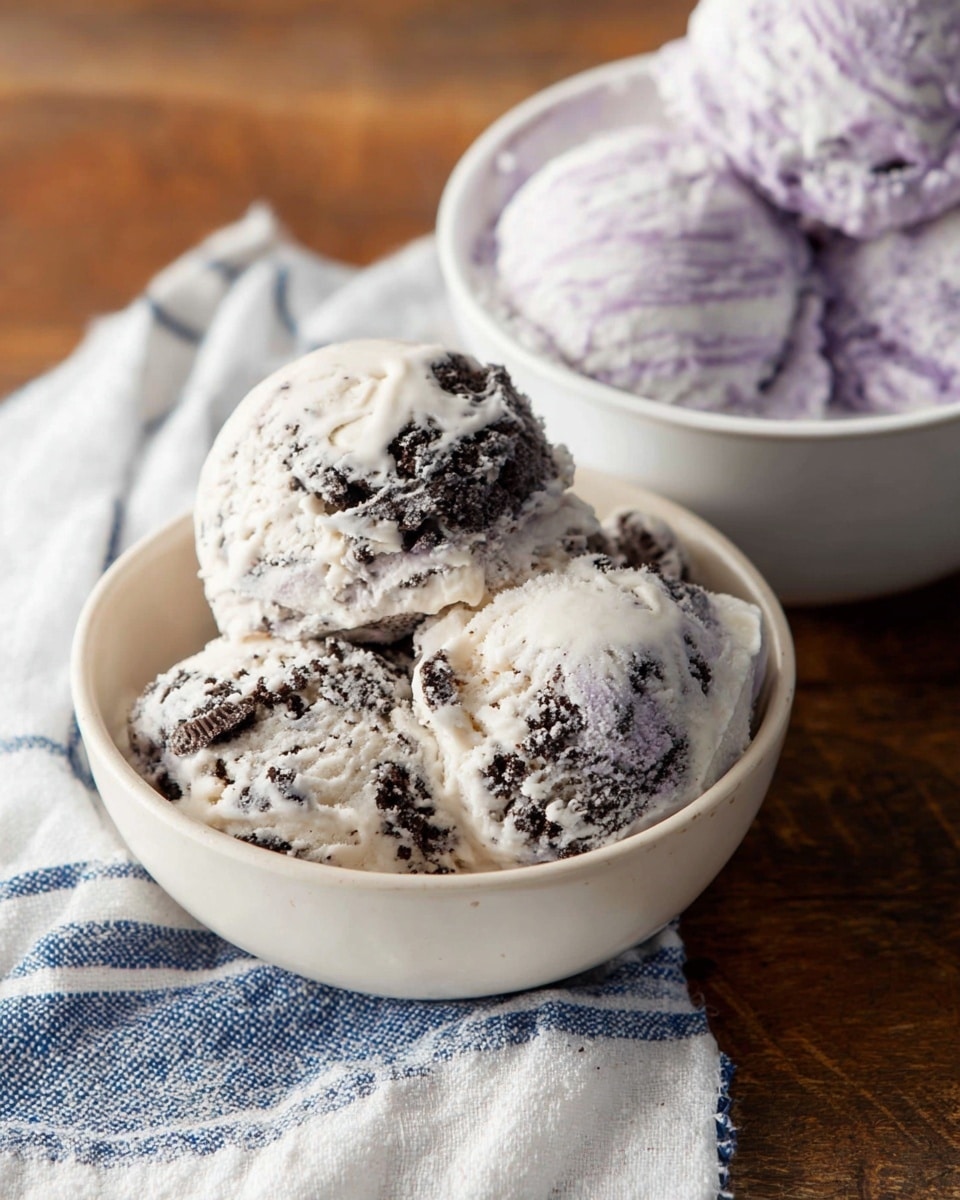 A close-up image showing two small ceramic white bowls filled with ice cream. The foreground bowl contains three scoops of cookies and cream ice cream, with a creamy white base and dark cookie bits swirled throughout, giving a marbled texture. The background bowl is filled with three scoops of light purple and white swirled ice cream, showing a soft and creamy texture. Both bowls rest on a wooden surface with a folded white cloth that has blue stripes beside them. The composition is simple and focuses on the cold, creamy texture of the ice creams. Photo taken with an iphone --ar 4:5 --v 7