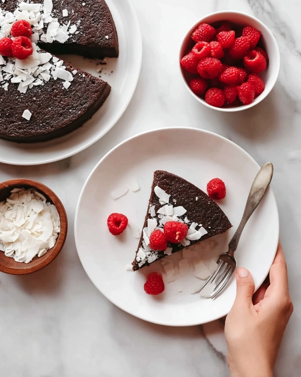A round dark brown cake is shown in two ways: one whole on a white plate and one slice on another white plate. The slice has three bright red raspberries and many thin white flakes, likely coconut, sprinkled on top and around it. A silver fork is placed beside the slice on the plate. A small white bowl filled with bright red raspberries and a wooden bowl with white flakes are near the plates. A woman's hand is holding the edge of the plate with the cake slice. The whole scene is set on a white marbled surface. Photo taken with an iphone --ar 4:5 --v 7