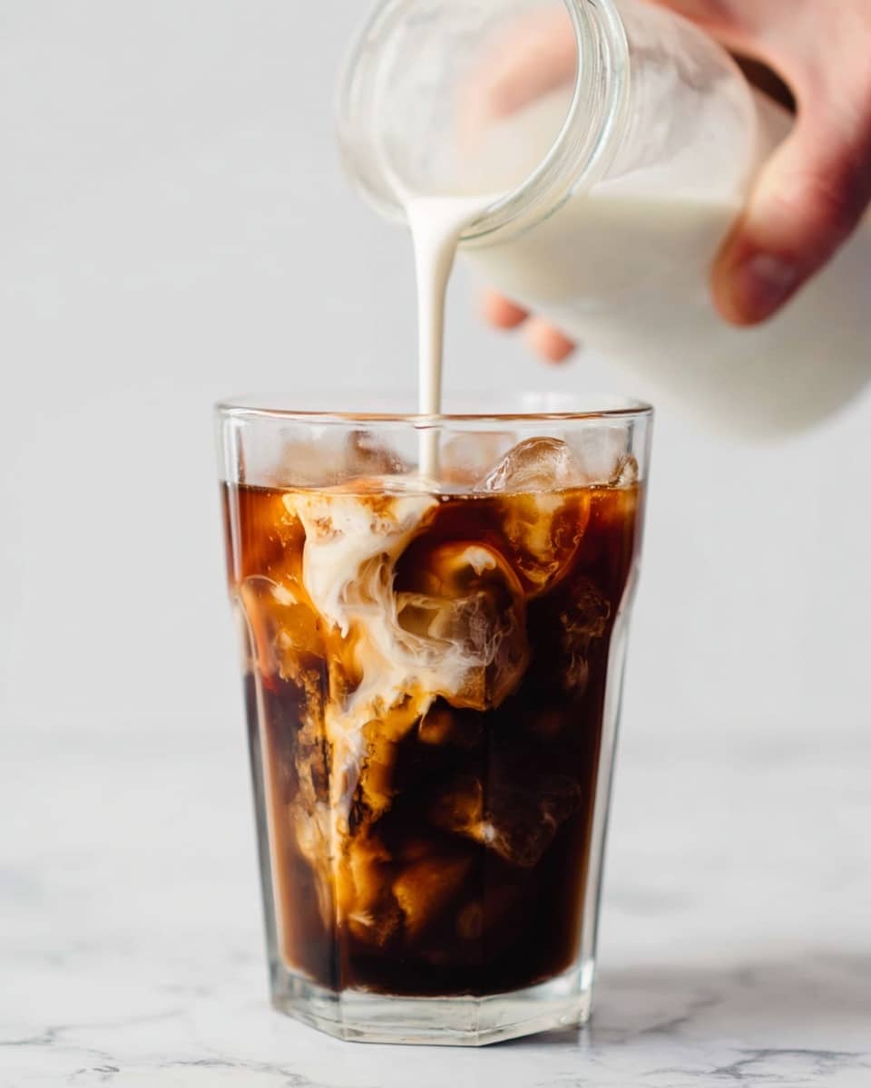 A clear glass filled with dark brown iced coffee and ice cubes showing different shades of brown inside, with cold cream being poured from a glass jar above; the cream creates white swirling patterns as it mixes into the coffee, all set against a white marbled background. A woman's hand is gently holding the jar, slightly blurred in the background. photo taken with an iphone --ar 4:5 --v 7