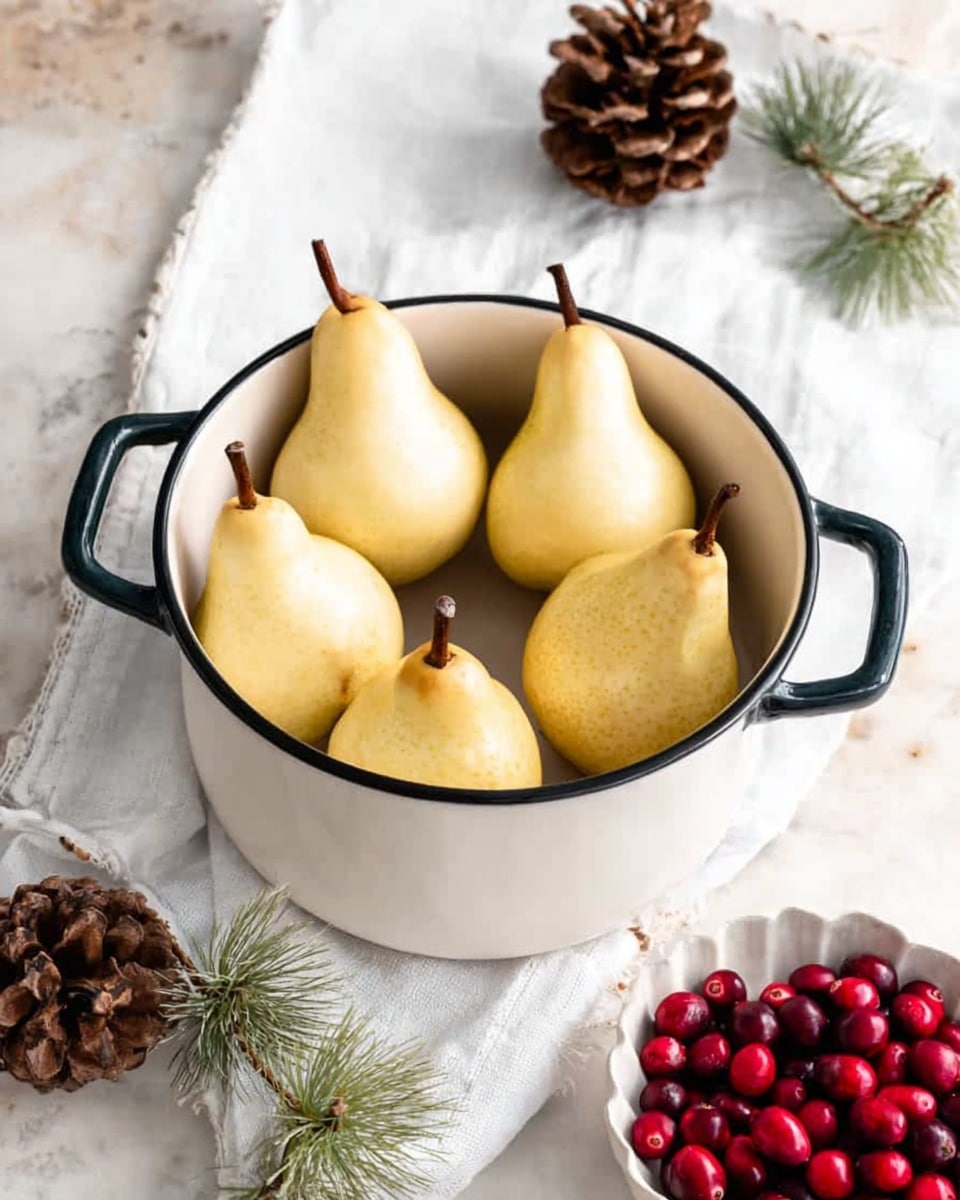 A round white pot with dark handles holds six peeled yellow pears standing upright inside. The pot is placed on a white marbled surface partly covered with a white cloth. Around the pot, there are a few pine cones and small green pine branches scattered, along with a white scalloped bowl filled with bright red cranberries. The light is soft, showing natural shadows. photo taken with an iphone --ar 4:5 --v 7