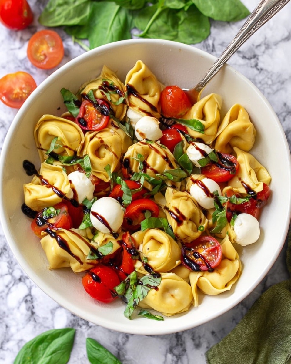 A close-up view of a bowl with light yellow tortellini pasta layered at the bottom, each piece showing smooth, folded edges. Scattered among the tortellini are halved bright red cherry tomatoes and small white mozzarella balls. Fresh green basil leaves are mixed evenly on top and around the pasta, adding a leafy texture. Dark brown balsamic glaze is drizzled over the entire dish in thin lines. The bowl is white with a rough brown rim and sits on a white marbled surface. photo taken with an iphone --ar 4:5 --v 7