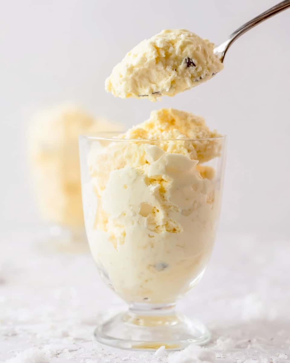 Three clear glass dessert cups are filled with a creamy, light yellow mixture that has small white marshmallow pieces and tiny nut bits visible, giving it a fluffy and chunky texture. In the background, a white bowl contains bright yellow pineapple chunks, slightly blurred but adding a pop of color. The dessert cups are placed on a white marbled surface, and a silver spoon lies in front of them, reflecting light softly. The setting is bright and clean, highlighting the creamy dessert's rich texture and pieces mixed inside. photo taken with an iphone --ar 4:5 --v 7