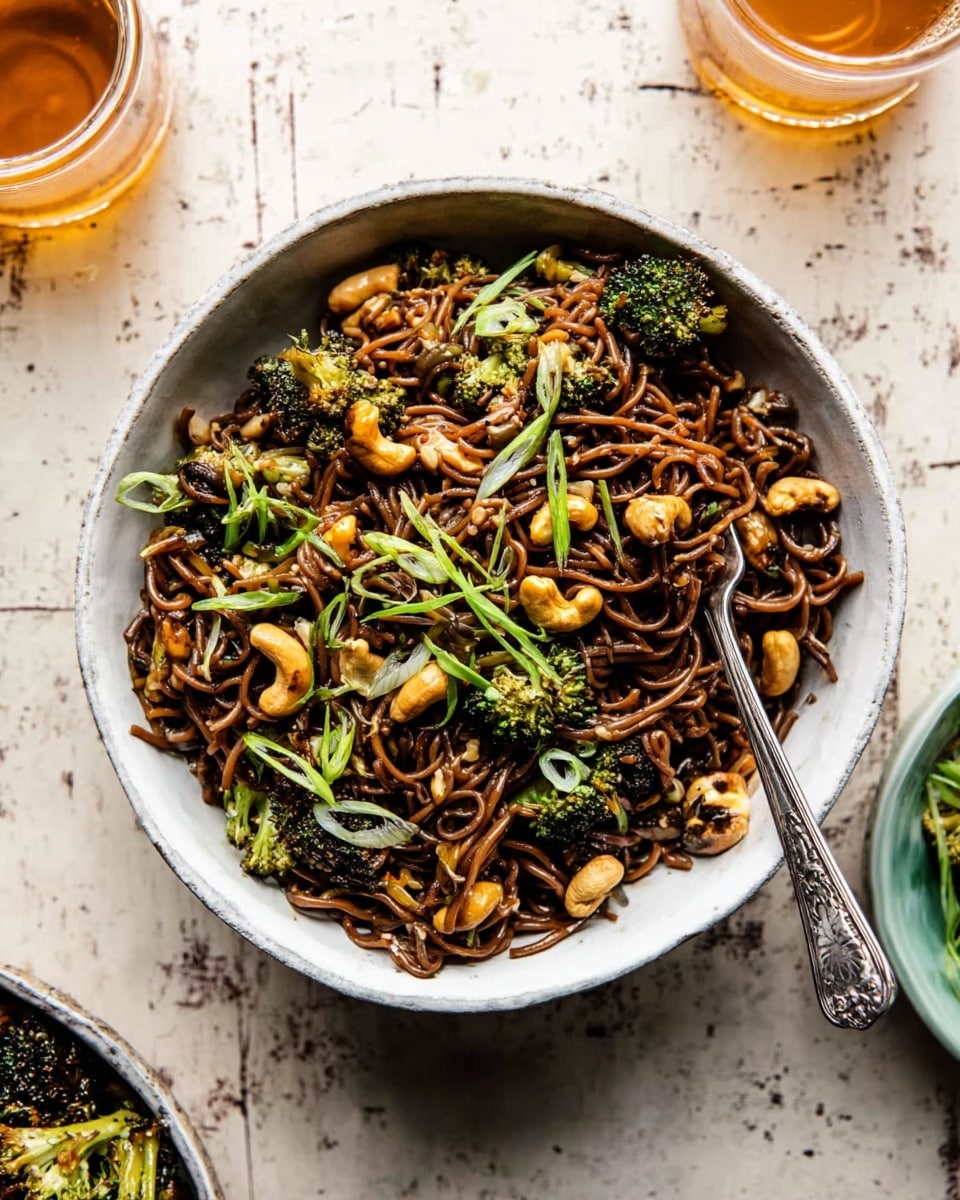 This image shows a close-up of a pan filled with cooked, wavy yellow noodles mixed with small, bright green broccoli pieces. On top of the noodles, there are clusters of dark brown and red crunchy chili flakes with sesame seeds. The noodles appear shiny and slightly oily, and the broccoli adds a fresh green contrast. The pan’s edge is visible along the bottom and sides, and the background is out of focus. photo taken with an iphone --ar 4:5 --v 7