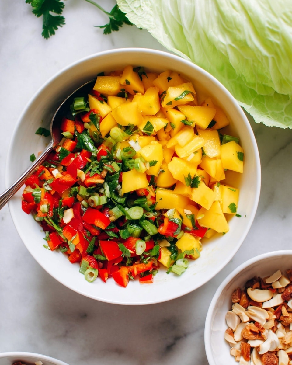 A white bowl filled with a colorful mango salsa, showing three layers of ingredients: the top layer has large yellow mango cubes, below are bright red bell pepper pieces, and mixed throughout are finely chopped green herbs and scallions. A silver spoon is partly visible inside the bowl on the left side. The bowl sits on a white marbled surface, and a large pale green cabbage leaf is placed near the top right corner. Part of another white bowl with light brown nuts and coconut flakes is seen at the bottom right corner. photo taken with an iphone --ar 4:5 --v 7