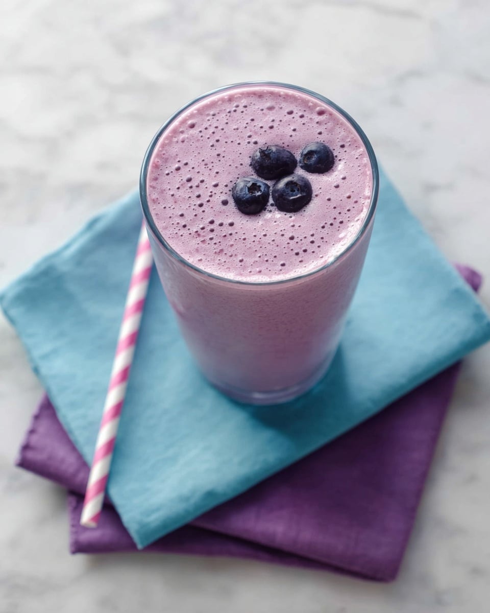 A tall clear glass fills with a smooth, light purple smoothie that shows tiny bubbles on the surface. Three small dark purple berries float on top, adding a bit of texture. The glass sits on two folded napkins, one pale blue and one bright purple, placed on a white marbled surface. Next to the glass is a white and pink striped straw, lying diagonally. The photo taken with an iphone --ar 4:5 --v 7