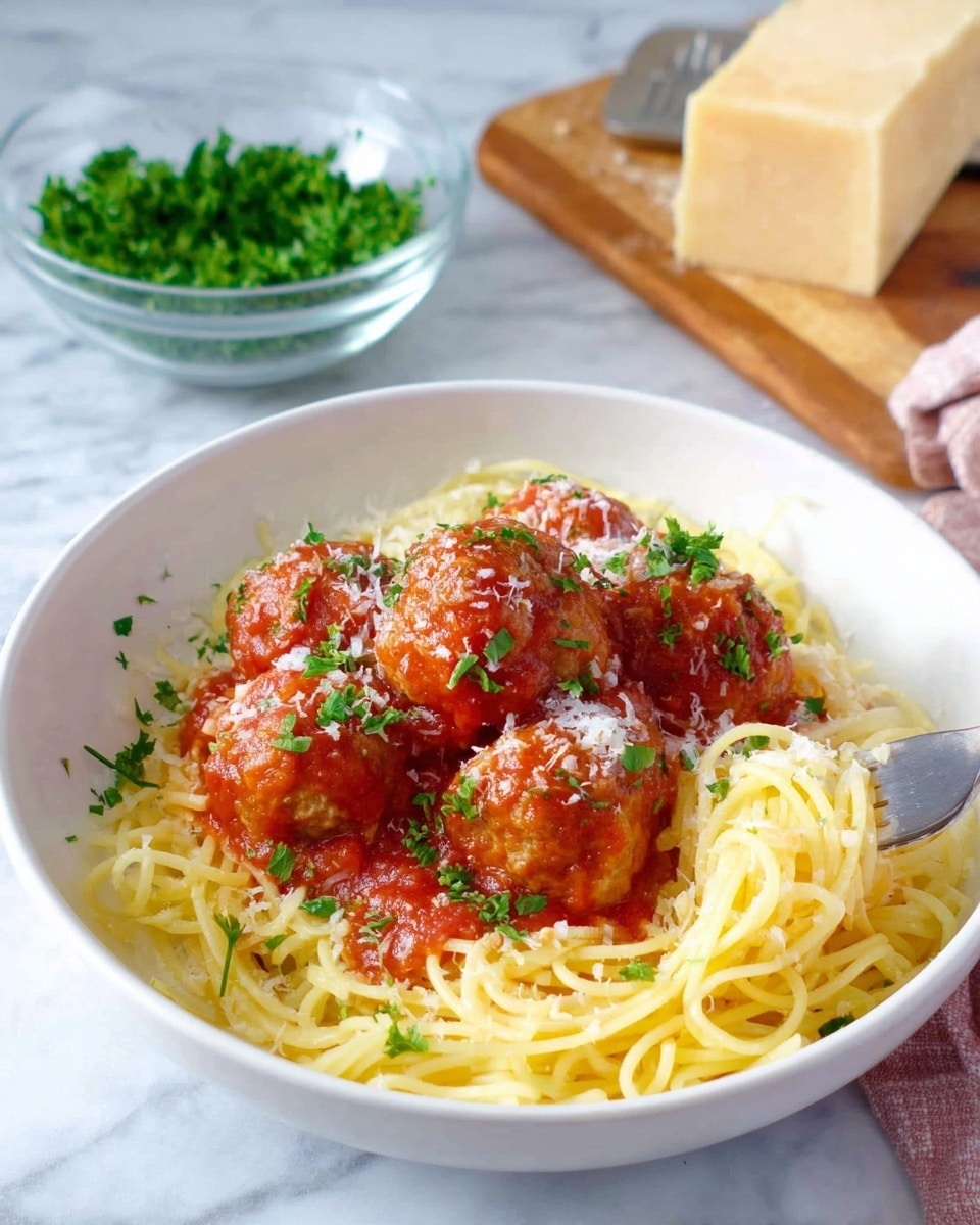 A white bowl filled with a base layer of plain yellow spaghetti noodles, topped with several medium-sized meatballs covered in a bright red tomato sauce with small bits of herbs. The meatballs are sprinkled with finely grated white cheese and chopped green parsley. In the background on a white marbled surface, a glass bowl contains more chopped green parsley, and a wooden board holds a round block of pale yellow cheese. A fork is placed inside the bowl on the right side, holding some noodles. photo taken with an iphone --ar 4:5 --v 7