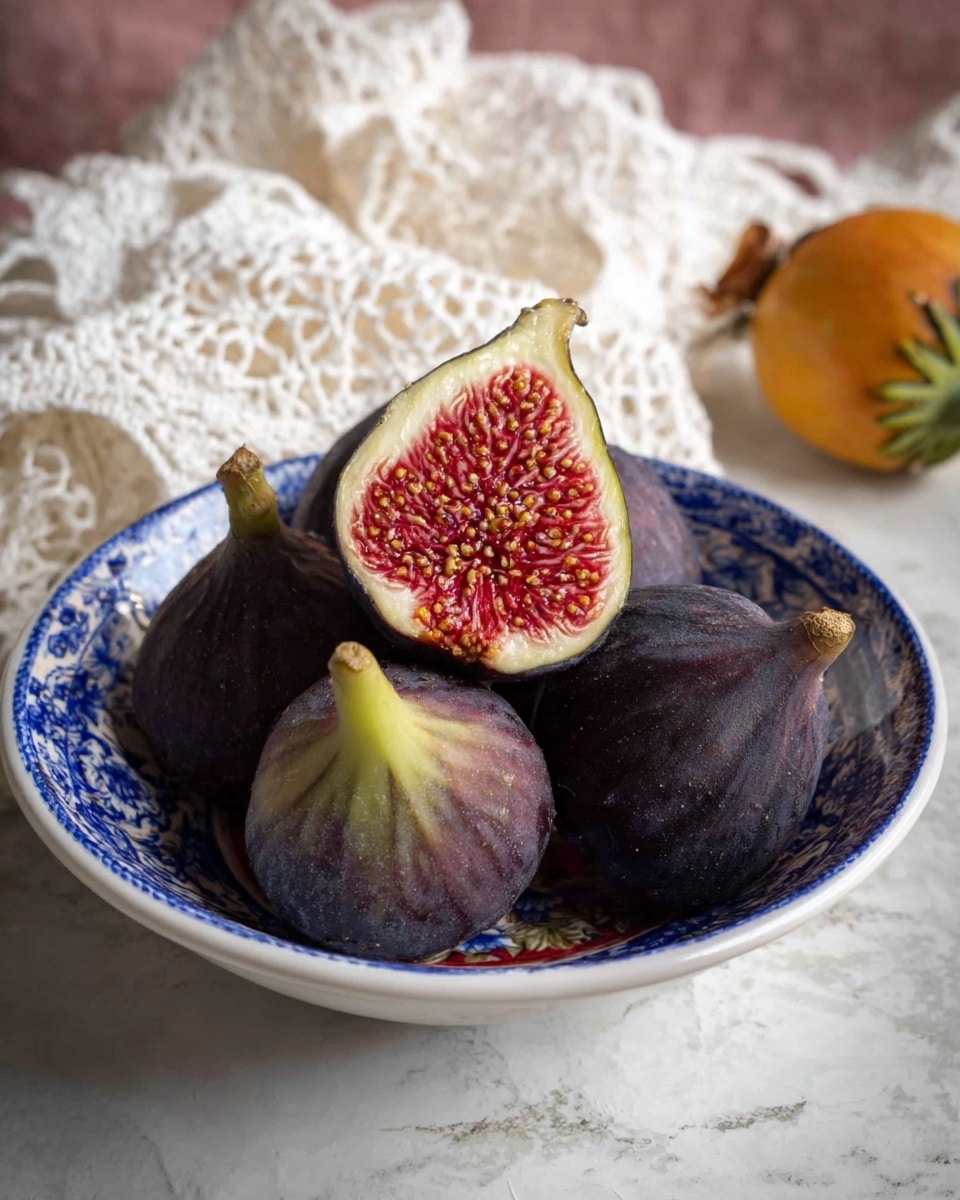 A bowl with a blue and white pattern holds five figs with dark purple and green skins. Four whole figs sit at the bottom, with one fig cut in half placed on top showing bright red and textured inside filled with small seeds. The bowl rests on a white marbled surface and there is a white crochet cloth in the background along with a small yellow-orange fruit with a green husk nearby. Photo taken with an iphone --ar 4:5 --v 7