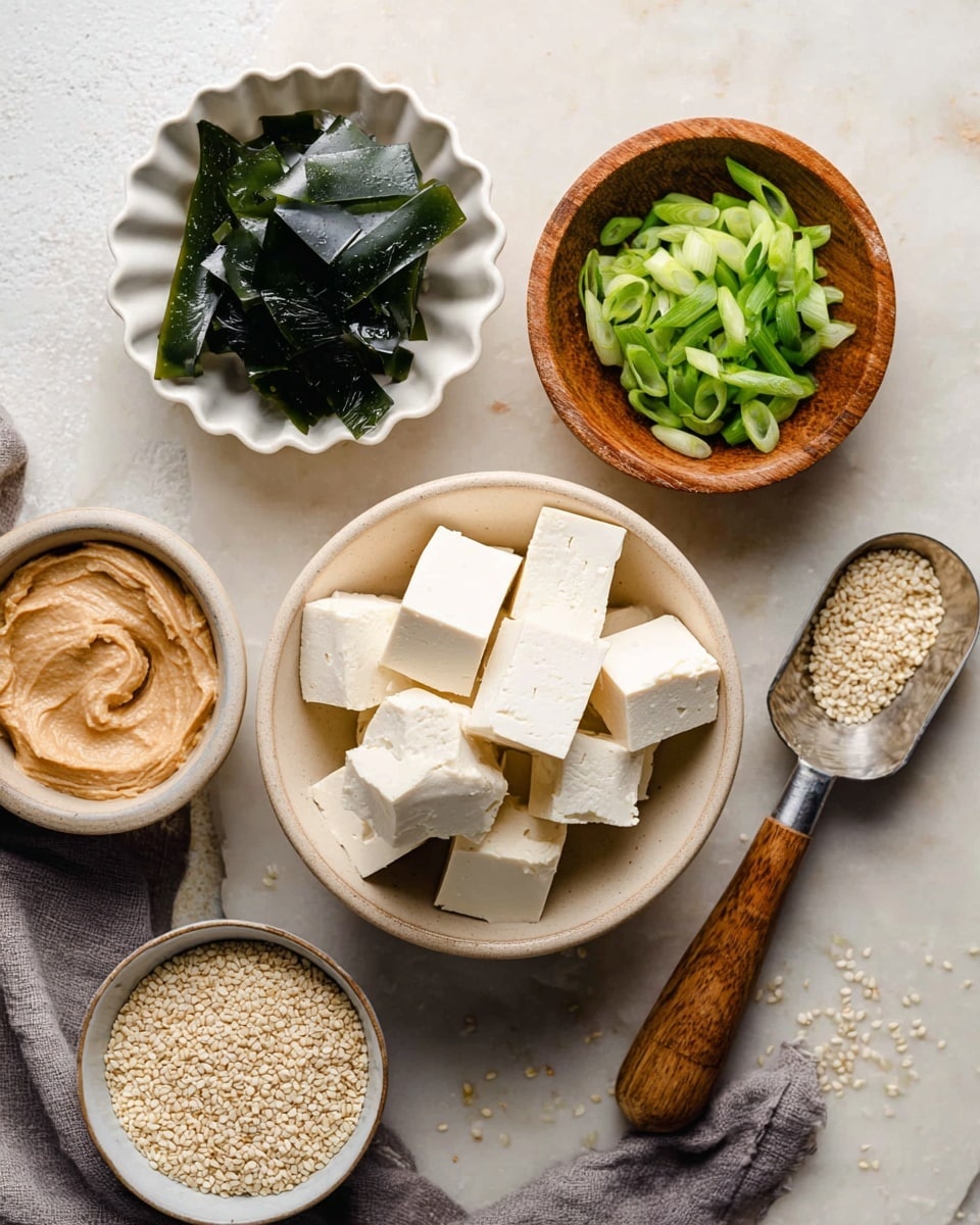 The image shows five small containers on a white marbled surface. At the center is a light beige bowl filled with white tofu cubes stacked in two layers. On the top left is a white scalloped bowl holding dark green, thin strips of seaweed with a slightly shiny texture. To the right of the seaweed is a small wooden bowl filled with bright green sliced scallions, layered loosely. Below the wooden bowl, a metal scoop with a wooden handle rests on the surface, filled with a smooth, light brown paste. Finally, at the bottom left, a small white bowl contains toasted white sesame seeds, some scattered on the surface nearby. A gray cloth is partly visible under the tofu bowl, photo taken with an iphone --ar 4:5 --v 7