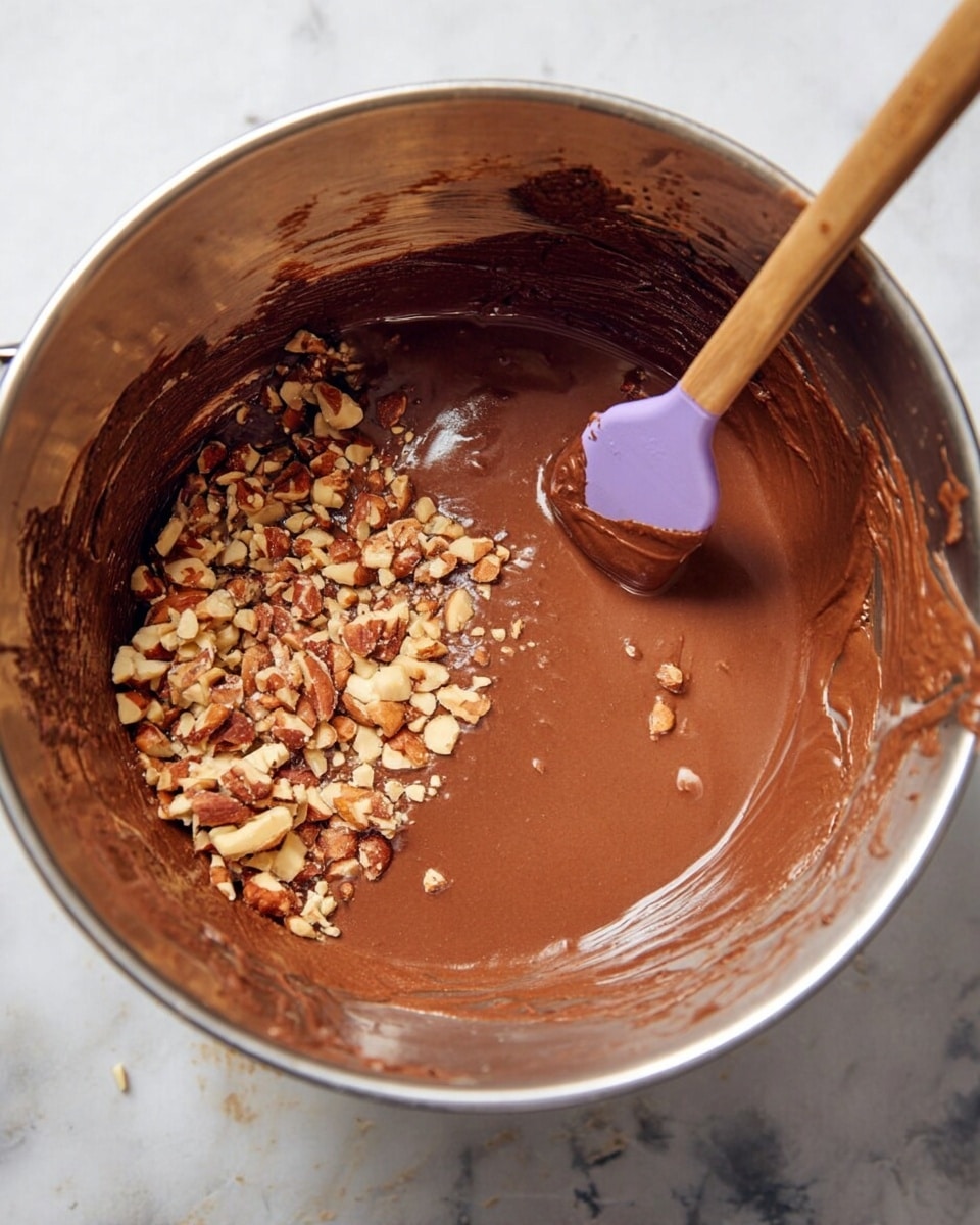 A silver mixing bowl holds rich brown chocolate batter with a smooth and creamy texture. On the left side of the batter, there is a pile of chopped light to dark brown nuts with rough, uneven edges. A wooden spatula with a purple silicone head is partially dipped into the batter on the right side, with batter coating its surface. The bowl sits on a white marbled surface, and some batter splatters cling around the inside edges of the bowl. photo taken with an iphone --ar 4:5 --v 7