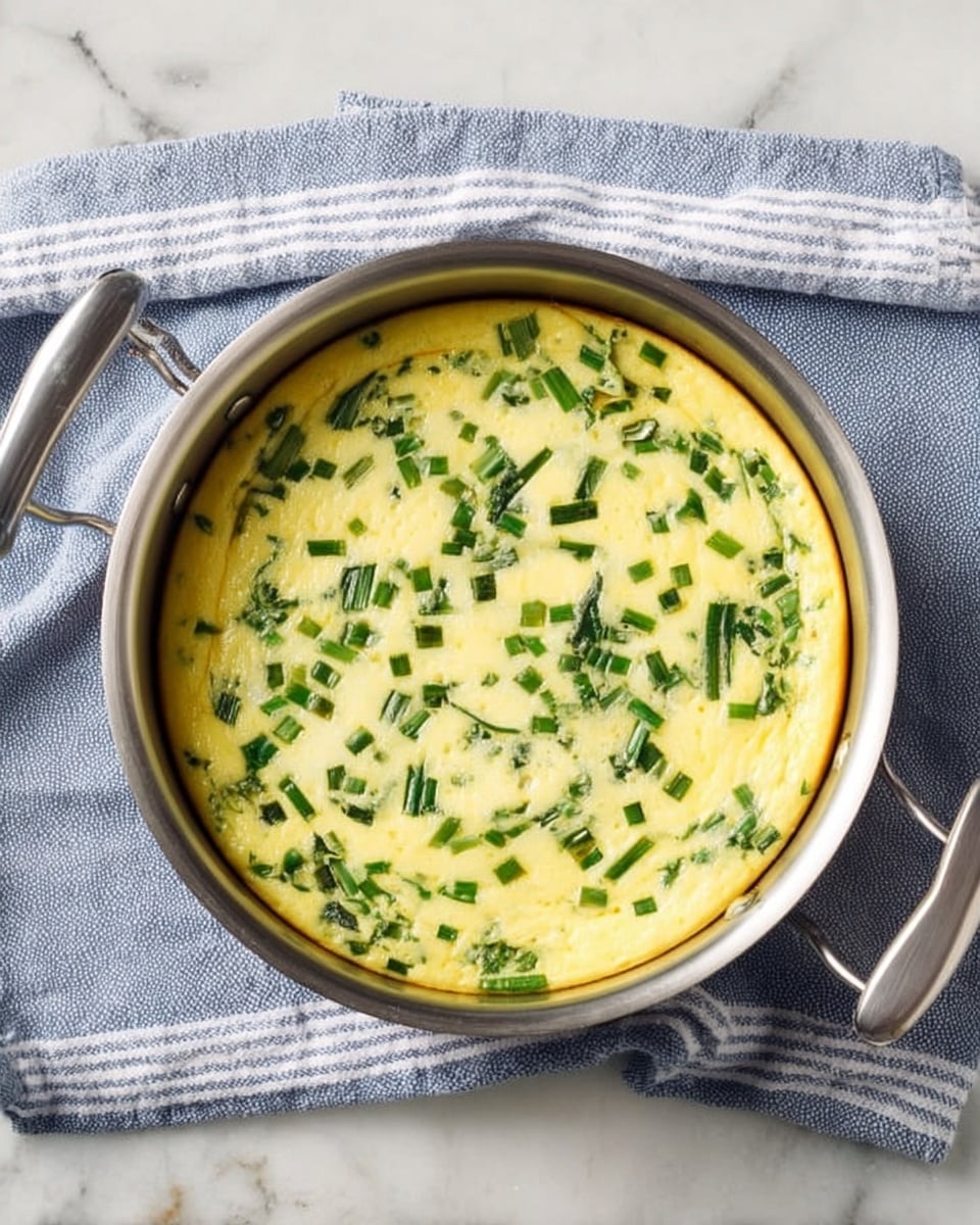 The image shows a round, light yellow frittata with small green vegetable pieces mixed evenly on the top layer. It is cooked in a silver metal pan with two thin handles on each side. The pan rests on a blue and white striped cloth, which lies on a white marbled surface. The frittata has a smooth texture with green herbs or vegetables giving a speckled effect across the surface. Photo taken with an iphone --ar 4:5 --v 7