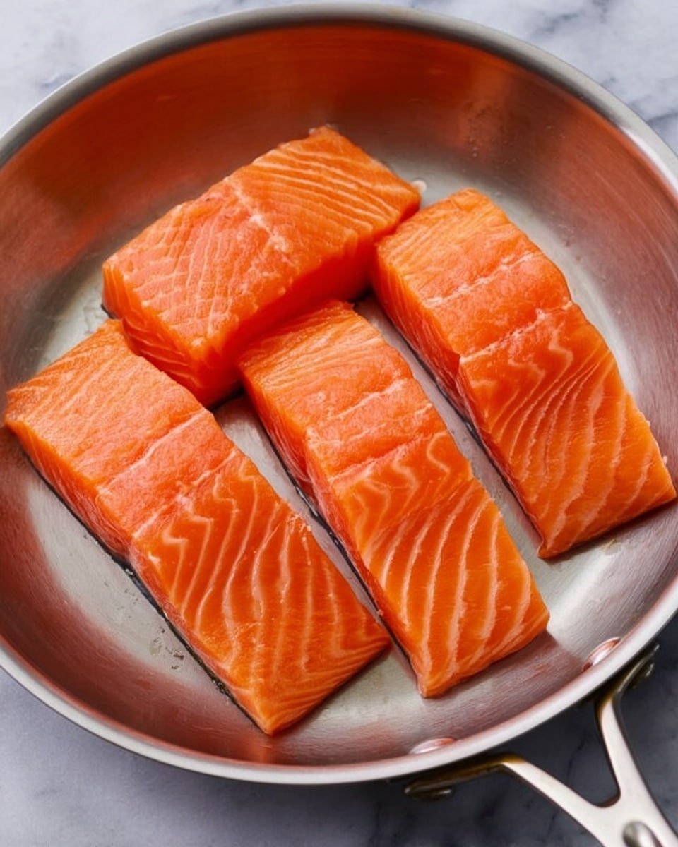 The image shows a close-up of four raw salmon fillets placed side by side inside a shiny silver frying pan. Each fillet has a bright orange color with visible white lines running through the flesh, showing the texture of the fish. The pan sits on a white marbled surface with soft lighting highlighting the freshness and smooth texture of the salmon pieces. photo taken with an iphone --ar 4:5 --v 7