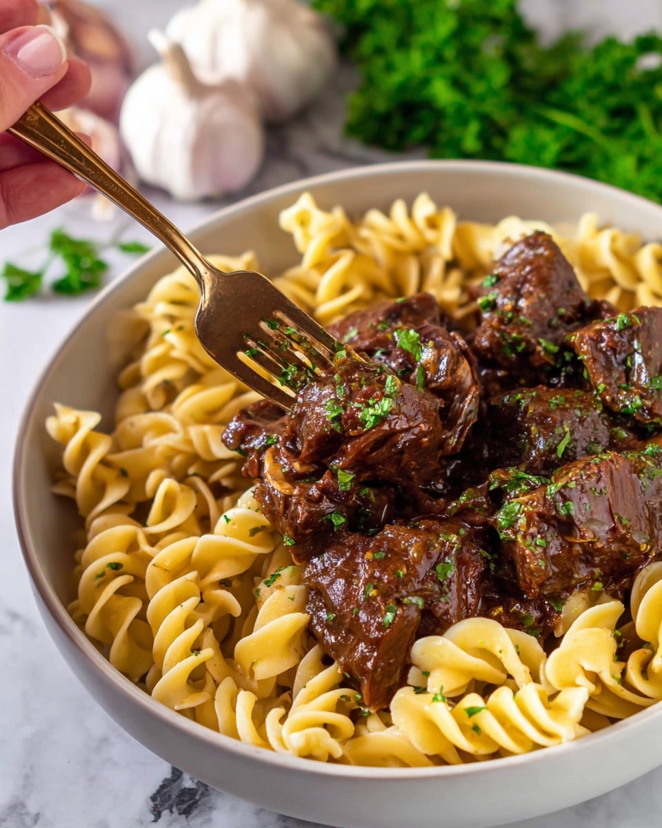 A white bowl filled with three layers: at the bottom is curly cooked yellow pasta, on top are large pieces of dark brown meat with a shiny sauce, and sprinkled green herb bits over both. A woman's hand is holding a fork stabbing one piece of meat. In the background, there are two whole garlic bulbs on a white marbled surface with some green parsley leaves peeking into the bottom right corner. Photo taken with an iphone --ar 4:5 --v 7