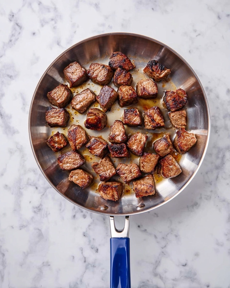 A top-down view of a round stainless steel frying pan with a blue handle placed on a white marbled surface, filled with evenly spaced, browned meat cubes showing a mix of golden and dark brown sear marks, each piece roughly the same size with a juicy texture visible, filling the pan without overlapping, and slight glistening oil spots around the meat. photo taken with an iphone --ar 4:5 --v 7
