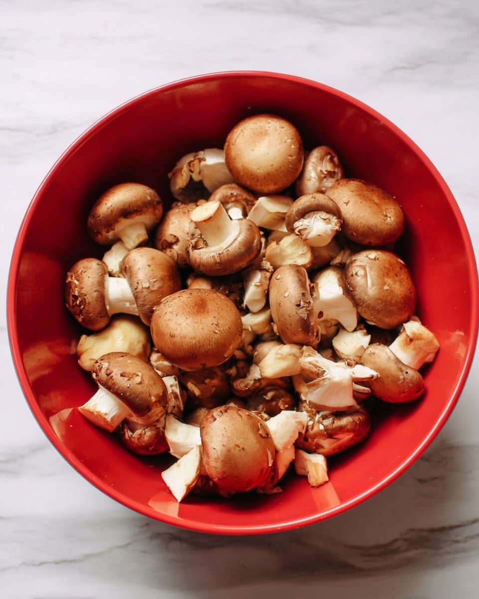A red bowl filled with whole and chopped brown mushrooms sitting on a white marbled surface. The mushrooms have light brown caps with some white stems showing where they are chopped. The bowl is round and deep, with the mushrooms loosely piled inside, showing a mix of whole pieces and smaller chunks. photo taken with an iphone --ar 4:5 --v 7