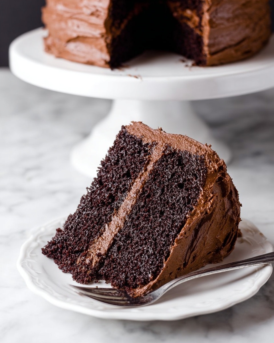 A thick slice of two-layer chocolate cake sits on a white plate with a small fork beside it. Each layer of the cake is dark, moist, and textured with tiny holes, separated and covered by a rich, light brown chocolate frosting that looks smooth and creamy. The frosting also covers the entire outer surface of the slice, creating a fluffy, thick coat. The background is a white marbled texture, and behind the plate, a white cake stand holds the rest of the cake. Photo taken with an iphone --ar 4:5 --v 7